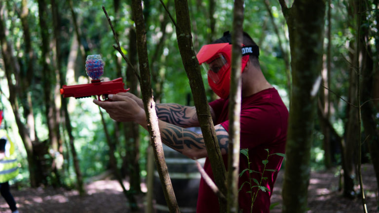 Person playing paintball in forest, aiming a marker through trees.