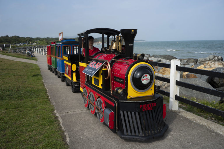 Miniature train ride along seaside path with colourful carriages and ocean view.