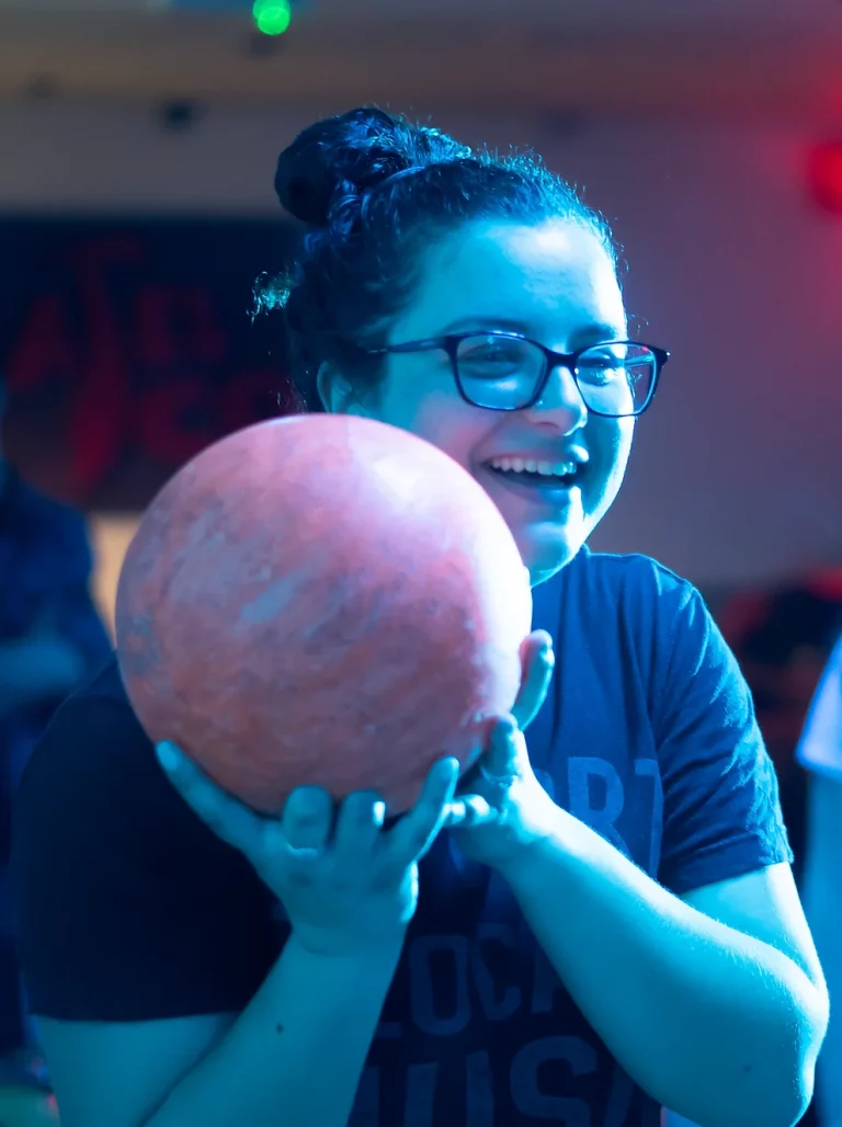 Smiling person with glasses holding a bowling ball in vibrant lighting.