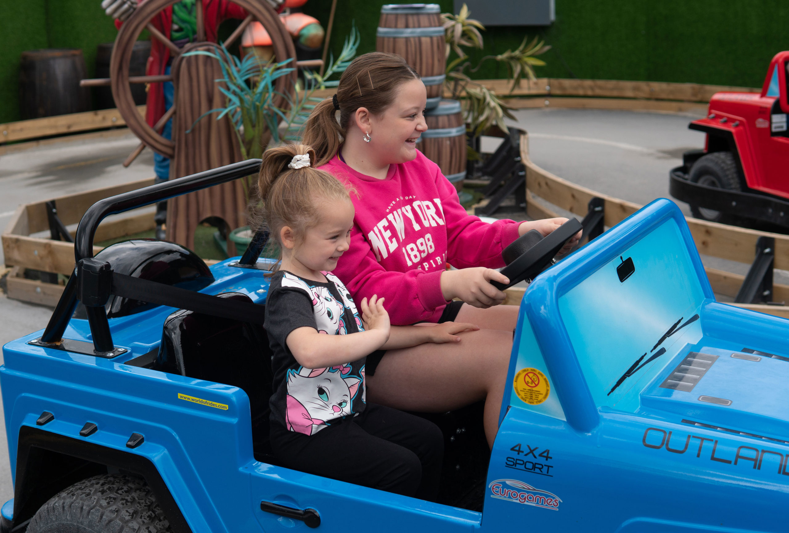 Two happy girls driving a blue toy car at an amusement park.