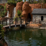 Miniature bridge over pond, waterfall, and stone house in quaint garden setting.