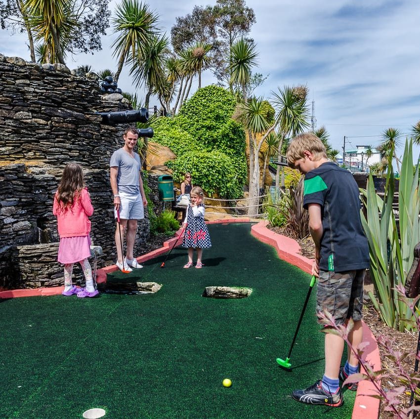 Family enjoys mini golf on a sunny day surrounded by palm trees.