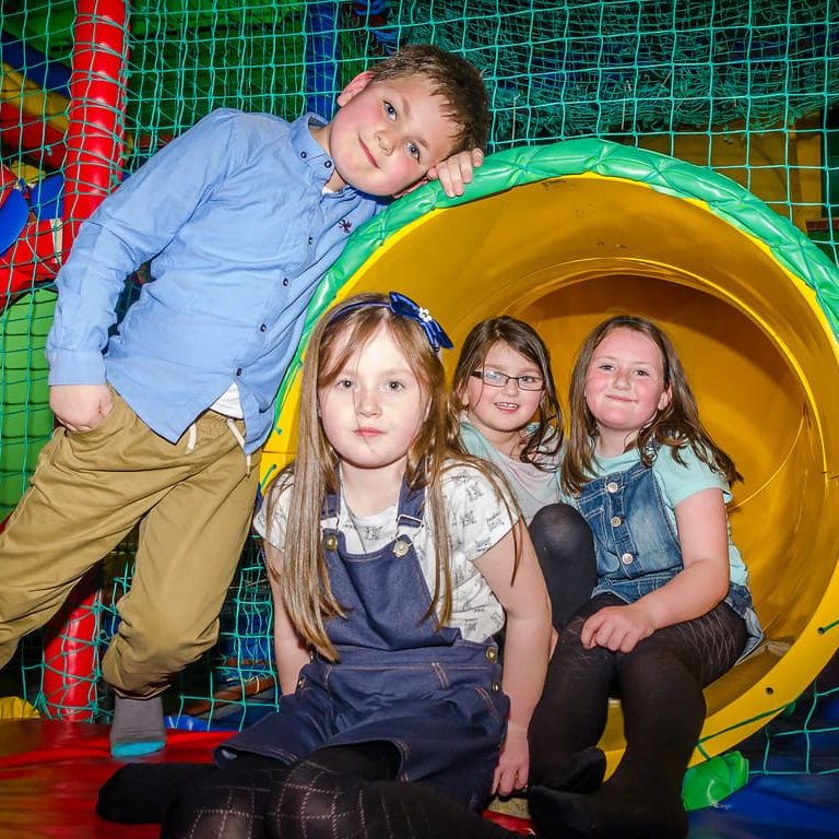 Children playing in indoor soft play area with colourful slides and nets.