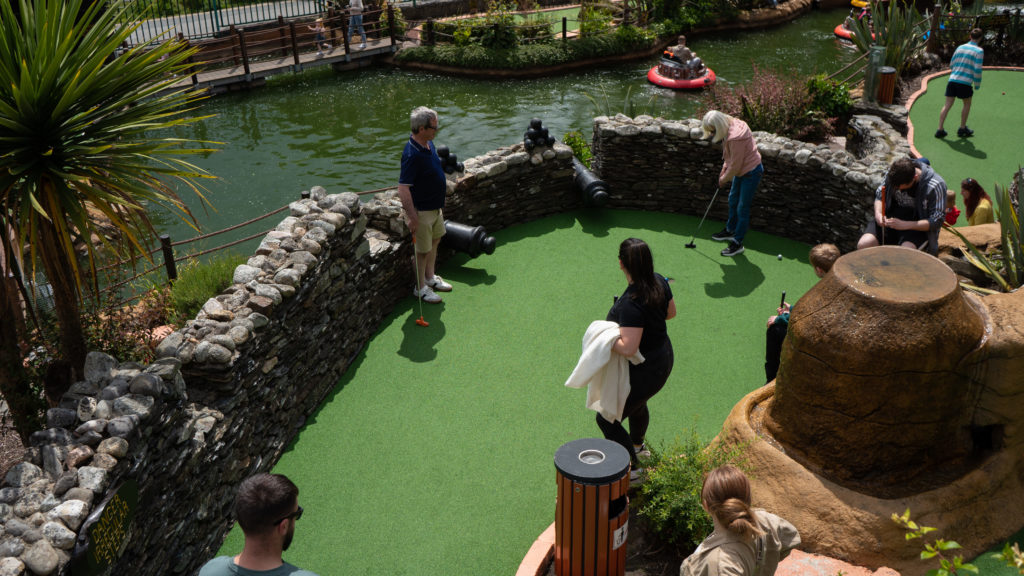 Mini golf course by pond with people playing under sunny skies.