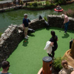 Mini golf course by pond with people playing under sunny skies.