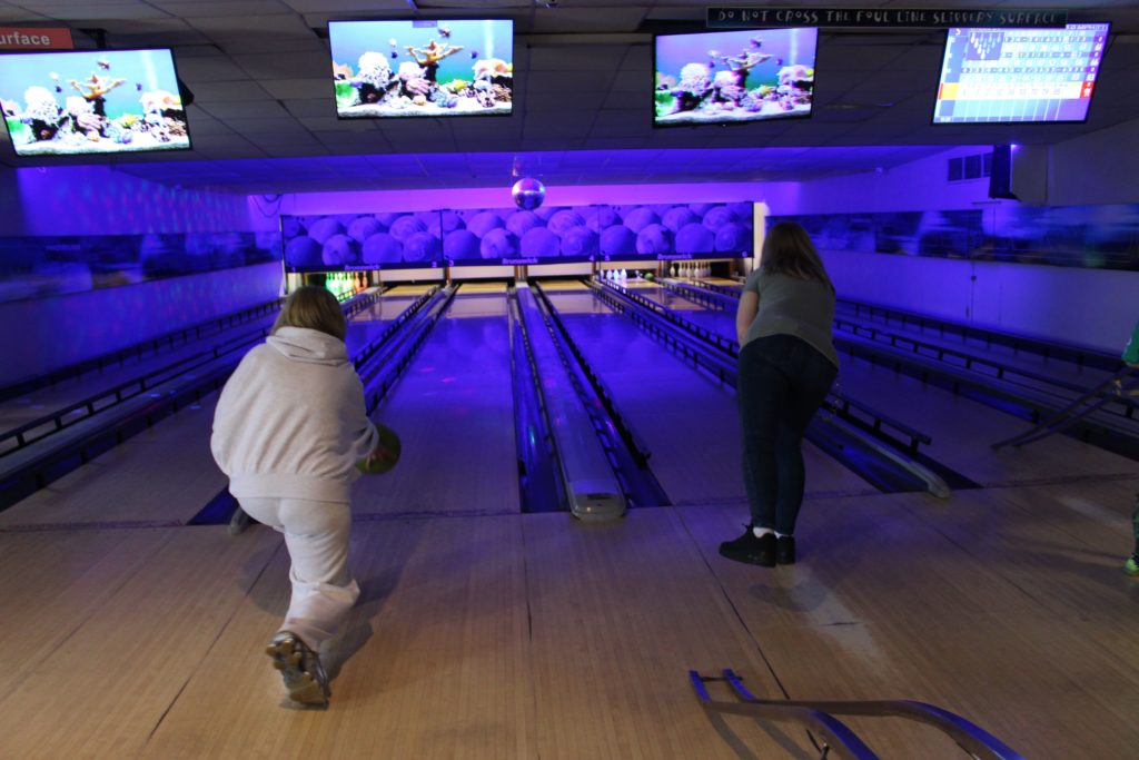 Two people bowling in a neon-lit alley, TV screens above lanes.